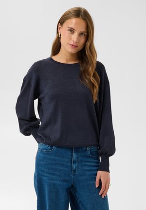 Jeune femme aux longs cheveux ondulés portant un pull bleu marine foncé et un jean bleu, debout, une main sur la hanche, sur un fond uni.