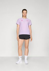 Világoslila sportos rövid ujjú póló rózsaszín logóval, fekete rövidnadrággal párosítva. A modell fehér tornacipőt és zoknit visel.