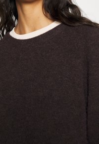 Dunkelbrauner Pullover aus einem strukturierten, weichen Stoff. Mit einem weißen Rundhalsausschnitt. Nahaufnahme, die die Details des Ausschnitts und der Schultern hervorhebt.