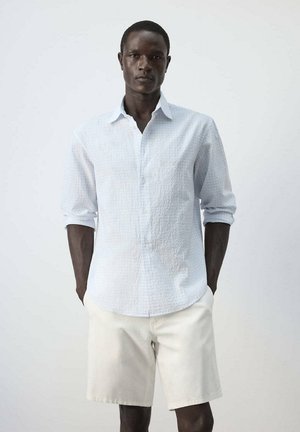 Hombre vistiendo una camisa blanca texturizada de manga larga con las mangas remangadas y pantalones cortos color crema, de pie frente a un fondo liso.