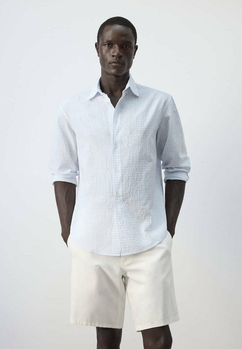 Homme portant une chemise blanche texturée à manches longues retroussées et un short couleur crème, debout devant un fond uni.
