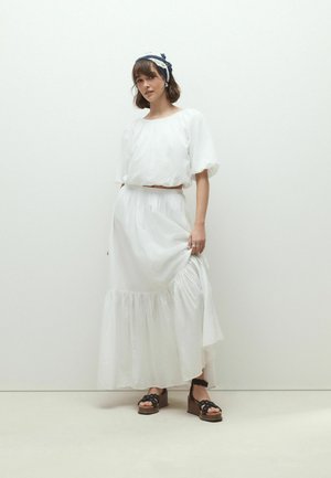 Nő áll egyszínű háttér előtt, fehér, puffos ujjú felsőt és hosszú, gyűrt szoknyát visel, fekete ék-nyeregszandált és kék-fehér fejpántot.