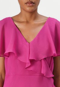 Robe fuchsia avec un décolleté en V et des manches à volants superposées, fabriquée en tissu léger avec une finition texturée.