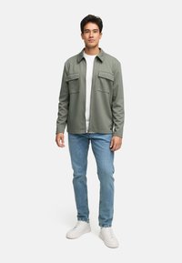 Grüne Jacke mit zwei Vorderaschen, getragen über einem weißen Hemd. Kombiniert mit hellblauen Jeans und weißen Sneakers. Einfaches Design und weiche Textur.