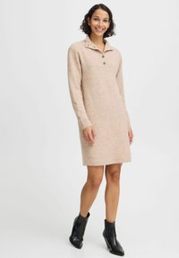 Gebreid jurkje in lichtbeige met een kraag en drie knopen. Heeft lange mouwen en een rechte snit, gecombineerd met zwarte enkellaarsjes.