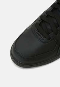 Gros plan sur la pointe d'une basket en cuir noir avec des lacets noirs et des détails perforés sur fond blanc.