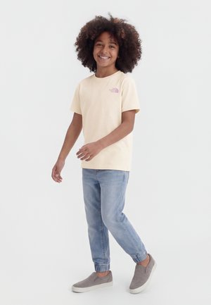 Enfant aux cheveux bouclés souriant, portant un T-shirt beige à manches courtes, un jean bleu clair avec des poignets élastiques, et des chaussures grises à enfiler.