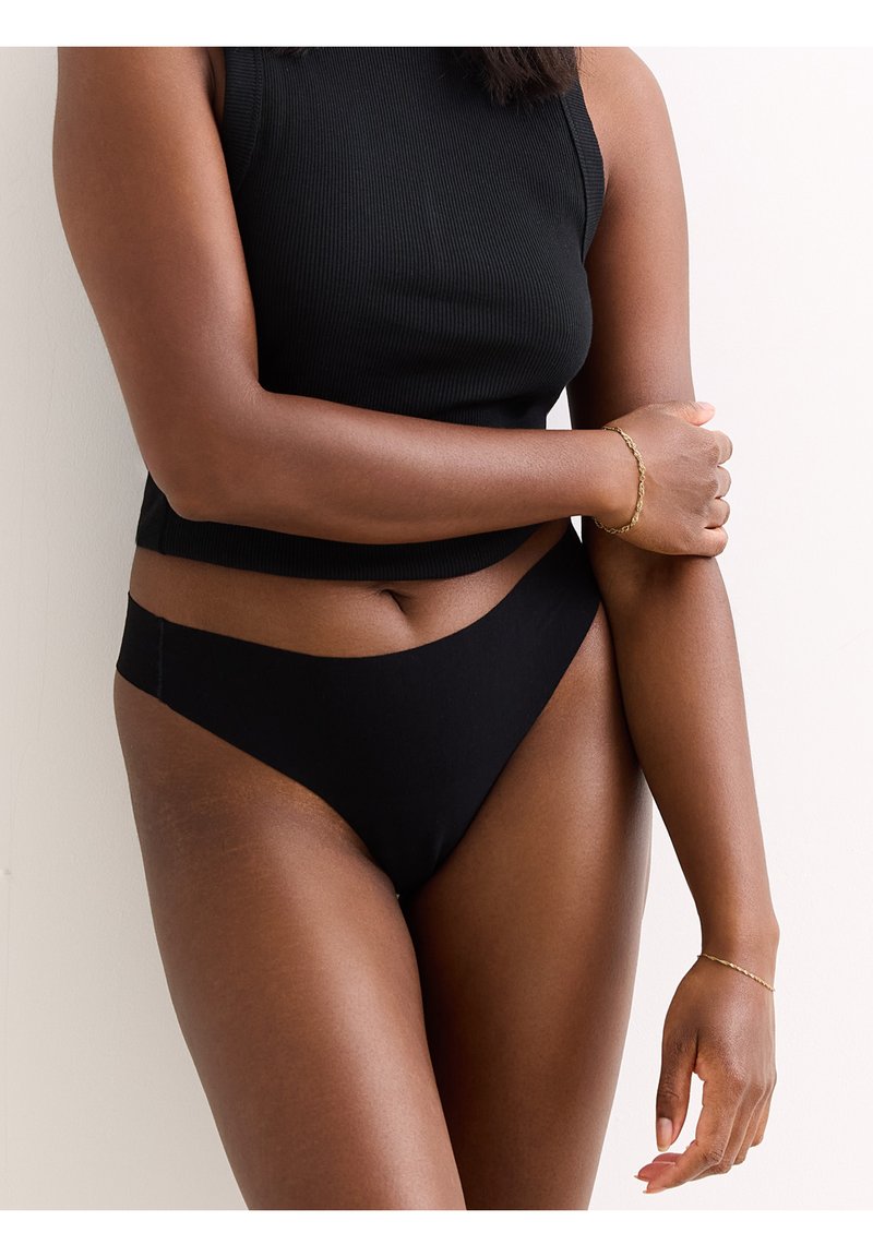 Schwarzes geripptes Crop-Top und passende Bikinihose. Der Stoff wirkt weich mit einem nahtlosen, tief sitzenden Design. Akzentuiert durch ein schlankes goldenes Armband.