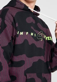 Černá mikina s kapucí, která má výrazný růžový pruhovaný vzor, texturovanou látku, přední kapsu a bílou tkanici. Zelený grafický text na hrudi.