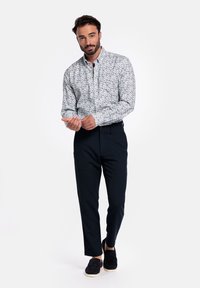 Witte overhemd met een multikleurig bloemenpatroon, zwarte broek en zwarte instappers, staand tegen een effen achtergrond.