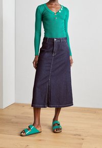 Mujer vistiendo una blusa verde de manga larga ajustada con botones dorados, una falda midi de mezclilla oscura con abertura frontal y sandalias verdes con hebilla.