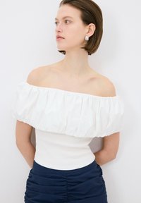 Top bianco off-the-shoulder con maniche a volantino, corpetto aderente e texture a coste, abbinato a una gonna blu navy arricciata. Design semplice ed elegante.