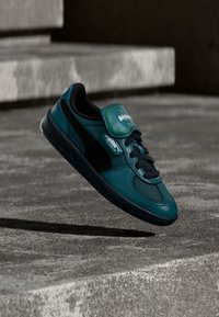 Teal leather sneaker with black accents, textured sides, lace closure, and a green branding tag on the tongue, floating above a concrete surface.