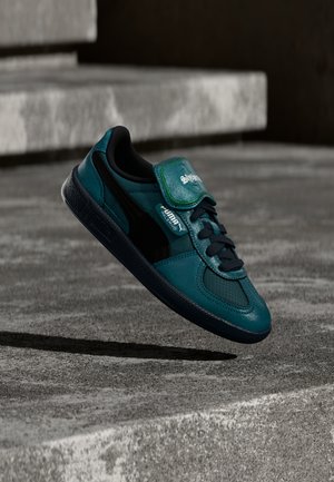 Turquoise leren sneaker met zwarte accenten, gestructureerde zijkanten, vetersluiting en een groen merkembleem op de tong, zwevend boven een betonnen oppervlak.