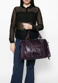 Sac à main en cuir violet avec deux poignées supérieures, comportant des accessoires en laiton, une poche frontale zippée et des détails de couture visibles.