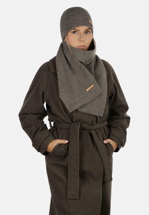 Frau trägt einen dunkelbraunen Mantel mit Gürtel, die Hände in den Taschen, eine graue gerippte Strickmütze und einen passenden langen Schal um den Hals gewickelt.