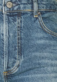 Gros plan sur la poche avant d'un jean en denim bleu avec un bouton en métal marqué "BOYISH JEANS" et des coutures jaunes visibles.