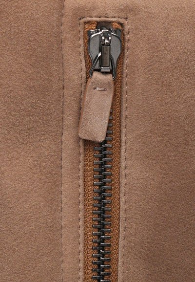 Közelkép egy barna velúr dzseki cipzárjáról, bőr húzófüllel és fém cipzárfogakkal, barna szövet háttéren.
