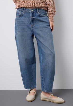 Donna che indossa jeans larghi di denim blu, un maglione corto marrone testurizzato e espadrillas Mary Jane beige, in piedi contro uno sfondo semplice.