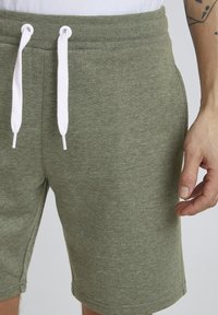Shorts en mélange de coton vert olive avec des cordons de serrage blancs, présentant une texture lisse et des poches latérales, vus de la taille vers le bas.