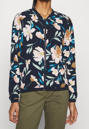 Chaqueta de cremallera azul marino con estampado floral en rosa, marrón y verde azulado, combinada con pantalones verdes oliva.