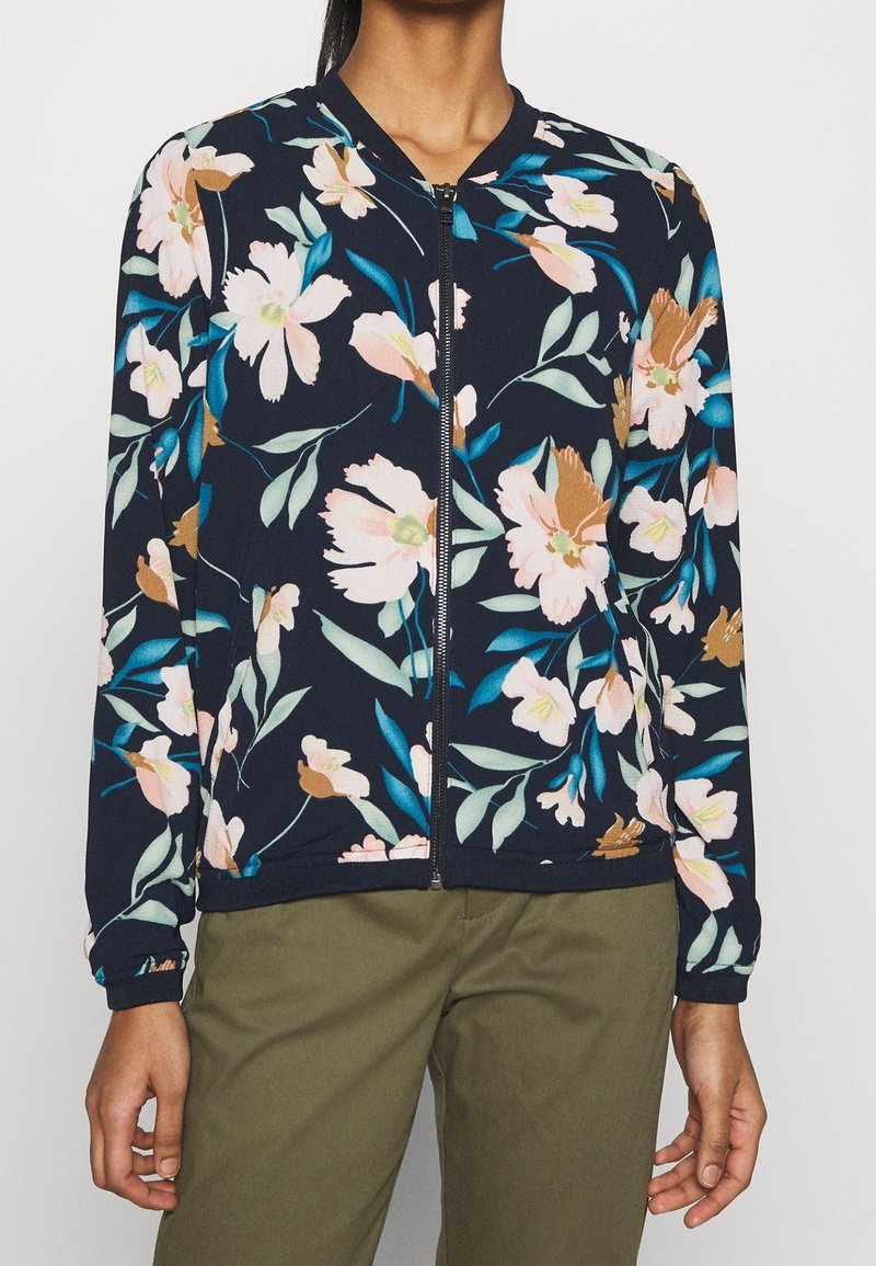 Mörkblå zip-up jacka med rosa, bruna och teal blommönster, bärs med olivgröna byxor.