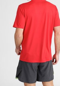 Hombre con una camiseta roja de manga corta lisa y pantalones cortos deportivos gris oscuro, de pie con la espalda hacia la cámara frente a un fondo claro.