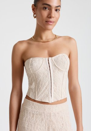 Donna che indossa un top corsetto in pizzo beige senza spalline e gonna a vita alta abbinata, con orecchini a cerchio dorati e una sottile collana d'oro.