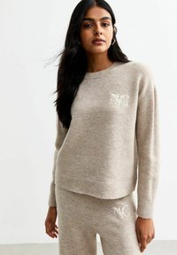 Pull en maille beige avec col rond, arborant un logo "NC" blanc sur la poitrine, associé à un pantalon beige assorti et à un design minimaliste.