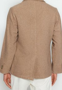 Blazer beige in lana con uno spacco sul retro indossato su pantaloni bianchi, che mostra la parte superiore della schiena e le braccia della persona.