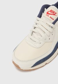 Nike Air Max kedai su sportu su kreminės spalvos odiniu viršumi, tamsiai mėlynomis detalėmis ir permatomu guminiu padu. Šnervės yra baltos su paminkštintu liežuviu.