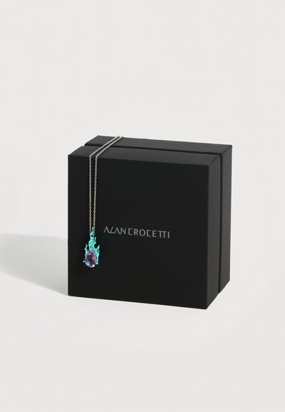 Μαύρο κουτί δώρου με ασημένιο λογότυπο, ασημένιο κολιέ με αλυσίδα που κρέμεται πάνω του, με μοτίβο δάκρυ μοβ με σχεδιασμό φλόγας σε τιρκουάζ.