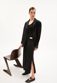 Zwarte op maat gemaakte blazer over een zwarte tanktop en rok met zijsplitten. Zwarte platte schoenen met een cirkelvormig detail. Bruine stoel op de achtergrond.