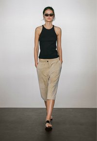 Černý top bez rukávů, béžové zkrácené kalhoty s kapsami a černé sandály. Modelka stojí proti světlému pozadí s neutrálními barvami.