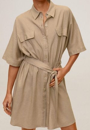 Vrouw draagt een beige jurk met knopen, korte mouwen, borstzakken en een gestrikte tailleband, staand tegen een effen achtergrond.