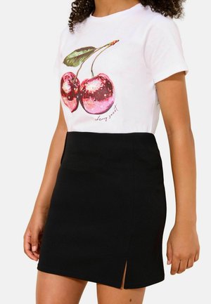 Nő, aki fehér pólót visel flitteres cseresznyékkel és „cherry sweet” felirattal, fekete miniszoknyával elöl hasítékkal párosítva.