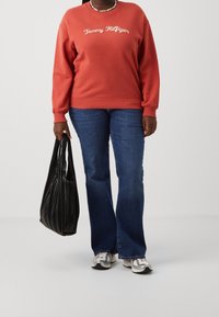 Personne portant un sweat-shirt rouge Tommy Hilfiger, un jeans bleu, des baskets, et tenant un sac à main plissé noir devant un fond uni.