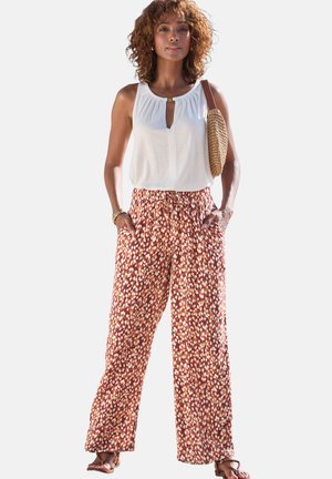 Frau trägt ärmellose weiße Bluse, rost- und weiß gepunktete Hose mit weitem Bein, trägt gewebte Schultertasche, steht mit den Händen in den Taschen.