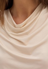 Krämfärgad topp med draperad halsringning, som har ett mjukt, slätt tyg och subtil struktur. Designen är enkel och åtsittande runt halsen.