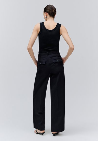 Femme avec les cheveux en chignon, portant des boucles d'oreilles argentées, un haut noir sans manches, un pantalon noir à jambes larges et des sandales noires à talons, vue de dos.