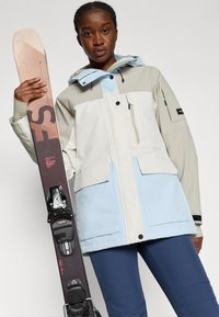 Hall hüppke hele sinine, beež ja hall suusajakk, millel on kapuuts, tõmblukkudega taskud ja reguleeritavad mansetid. Mudel hoiab suuska koos sidemete jaotusega.