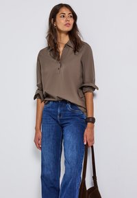 Hellbraunes, kragenloses Blusenshirt mit Knopfleiste und hochgekrempelten Ärmeln, kombiniert mit blauen, weit geschnittenen Jeans aus Denim und einer braunen Tasche. Glatte Stoffstruktur.