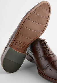 Brown leather dress shoe featuring brogue detailing, round toe, and a rubber sole with ridged traction patterns. Label reads "NEXT Motion Flex."