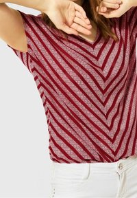 Top de manga corta a rayas rojas y blancas, con un diseño de cuello en V, fabricado en un tejido texturizado y con un patrón de flechas. Combinado con pantalones blancos.