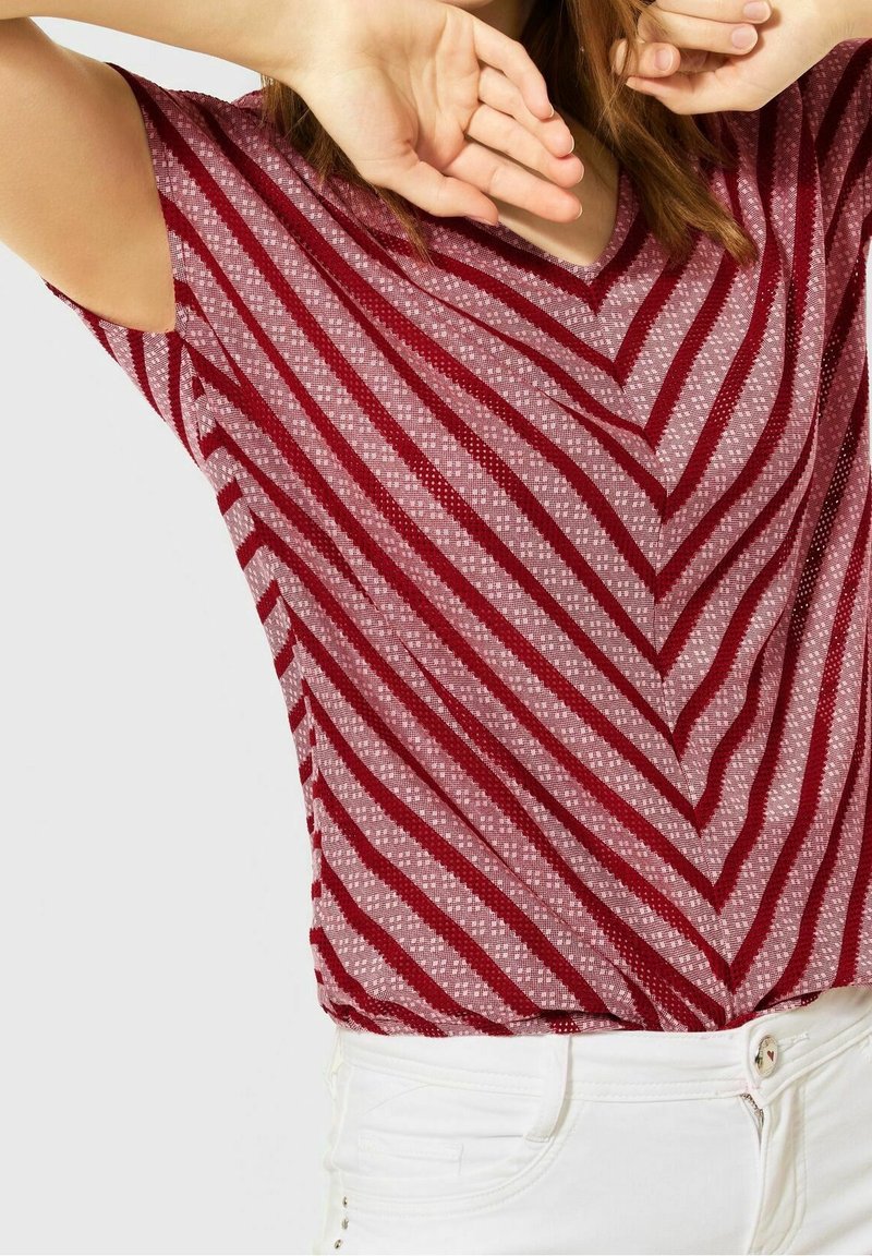 Top de manga corta a rayas rojas y blancas, con un diseño de cuello en V, fabricado en un tejido texturizado y con un patrón de flechas. Combinado con pantalones blancos.