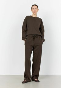 Hnědá mikina a odpovídající tepláky vyrobené z měkké látky, s kulatým výstřihem a elastickým pasem na šňůrku. Celý outfit doplňují ploché boty.