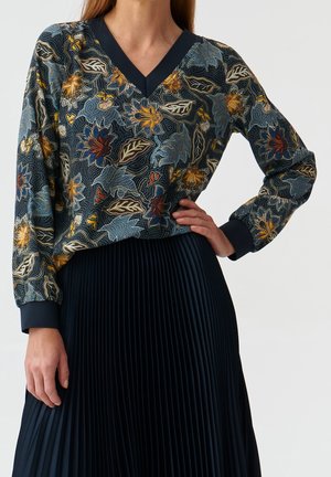Woman wearing a floral-patterned long-sleeve V-neck top with dark cuffs paired with a black pleated skirt, hand on hip.