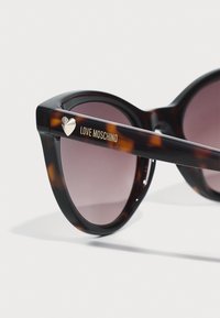 Silmany kehykset, joissa on pyöreät linssit, tummat gradienttilinssit, kultainen "Love Moschino" -brändäys ja sydänkoriste varressa.