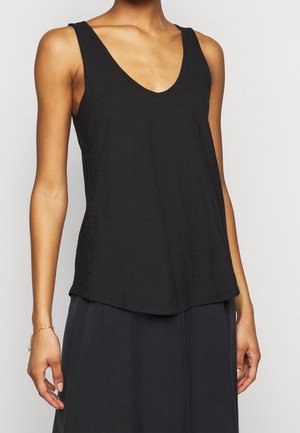 Persona delgada con una blusa negra sin mangas y escote en V, y una falda oscura, de pie frente a un fondo claro y sencillo.