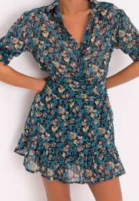 Kukkamekko tumman turkoosilla taustalla, jossa on lyhyet hihat, kauluksellinen kaula-aukko ja röyhelöinen helma. Materiaali on kevyt ja läpikuultava.